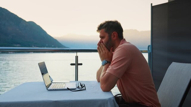 Stressed remote worker on balcony transitions to focused productivity while using laptop by tranquil lakeside at sunset with scenic mountain horizon view
