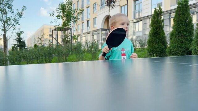 A cheerful young boy raises a ping pong paddle with an eager smile. He looks away playfully behind an outdoor table tennis table. Green trees and modern residential buildings surround him.