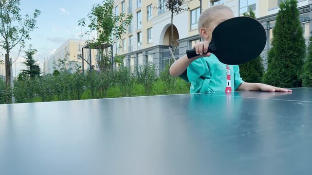 A happy young boy holds a ping pong paddle with excited wide eyes. He stands enthusiastically at an outdoor table tennis court. Modern buildings and trees surround the cheerful courtyard.