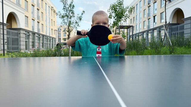 An excited young boy swings a table tennis paddle towards a yellow ping pong ball. He plays with intense joy at an outdoor table. Modern apartment buildings surround the active courtyard.