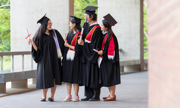 Group of asian university graduates talking together with diplomas on campus walkway, Cheerful graduates in academic gowns and caps holding degree scrolls standing and chatting on graduation day