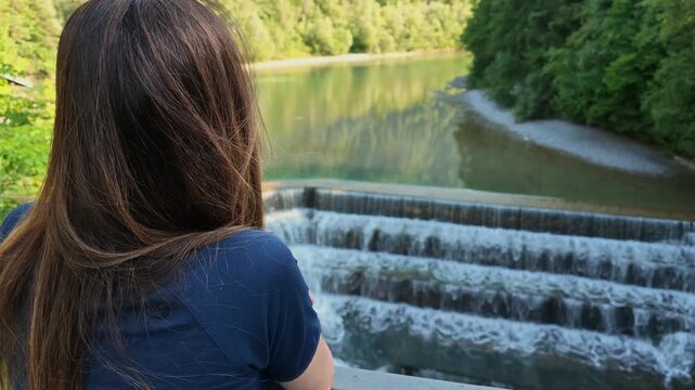 Young woman with long hair seen from behind as she admires the tiered Lechfall cascade and the turquoise Lech River from a viewpoint in Fussen, Bavaria.