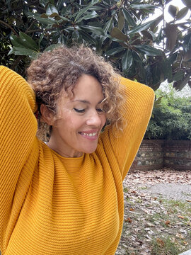 Curly-haired woman smiling, relaxing outdoors in a park, experiencing joy and freedom while stretching her arms above her head, surrounded by green foliage and fallen leaves