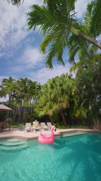 Vertical Screen: Woman relaxing by the pool on a yellow towel, a pink flamingo float on turquoise water. Tall palm trees, white clouds, sun loungers, umbrella and grill in a tropical backyard.