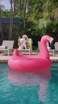 Vertical Screen: Woman relaxing by the pool tilts her head back to the sun on a lounger with a cocktail glass. Pink flamingo float on turquoise water, white hat, blue swimsuit, palms on a sunny day.