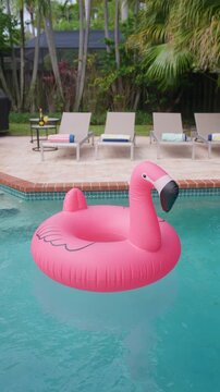 Vertical Screen: Pink inflatable flamingo float drifts on turquoise pool water. Sun loungers with rolled towels, bottle and cocktail on side table, palm trees behind. Tropical backyard summer vacation
