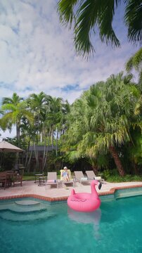 Vertical Screen: Woman in sun hat and blue swimsuit relaxes on a lounger with a drink by a turquoise backyard pool. Pink flamingo float, tall palm trees, patio umbrella, grill and cloudy blue sky.