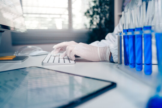 Laboratory scientist wearing protective gloves typing on a computer keyboard beside blue liquid samples in test tubes at a modern research laboratory.