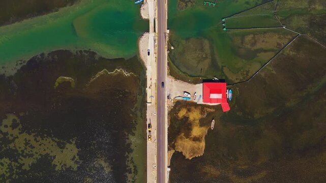 Amazing aerial top down view of a causeway dividing a marshy lagoon, with a red building and traditional fishing nets, while cars are driving on the road in Narta, Albania