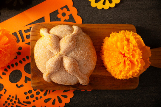 Mexican Pan de Muerto sweet bread on a wooden board, surrounded by cempasuchil marigold flowers and papel picado for Day of the Dead. It is a main element in the altars and offerings.