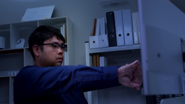 Asian businessman stretching arms while working late at office desk to relieve stress from overtime corporate project