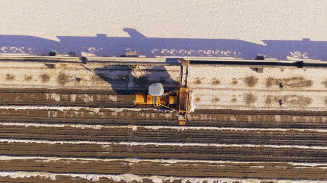 Aerial top-down view of a large composting machine on rails turning long windrows of organic matter at a farm facility during a sunny winter day, producing natural soil fertilizer