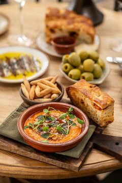 Gourmet Spanish tapas spread featuring red pepper dip in a terracotta cazuela, artisan focaccia bread, green olives, breadsticks, and white wine.