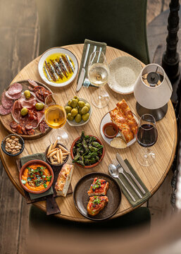 Top-down view of a round wooden table filled with Spanish tapas including charcuterie, olives, padron peppers, anchovies, focaccia, and white wine.