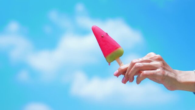 Close-up of hand holding melting watermelon popsicle dripping under bright blue summer sky. Hot weather, heatwave and refreshment lifestyle concept.