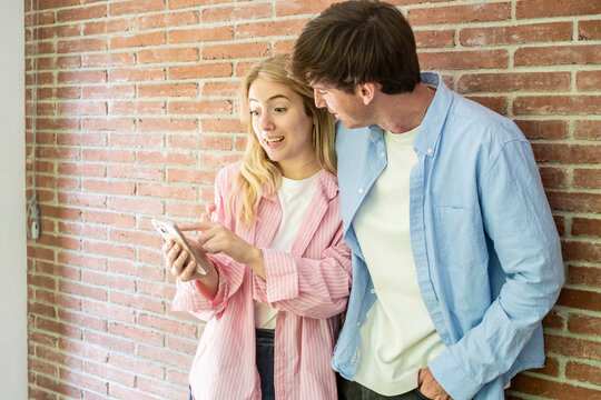 Young couple standing against a brick wall, sharing content on a smartphone, with the woman showing surprise and pointing to the screen, while the man leans in to see