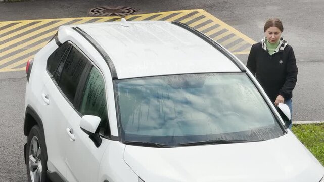 A thoughtful woman steps out of a white SUV on a damp paved parking lot. She appears serious and focused as she pauses beside her open vehicle.