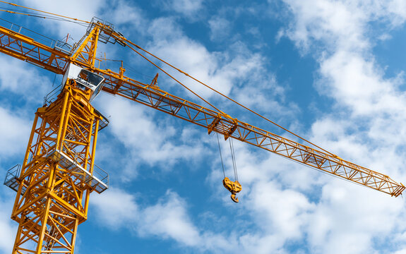 Large yellow construction crane jib extends toward blue sky with white clouds. Heavy industrial machinery uses steel lattice boom, lifting hook for building projects