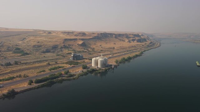 Grain silos and agricultural storage facility beside the Columbia River and Interstate 84 in Oregon, with wildfire smoke creating hazy conditions across the dry landscape.