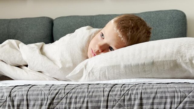 A young boy lies under a cozy blanket and watches tv on a soft sofa. He rests his head peacefully on a pillow. His quiet gaze shows calm and restful relaxation.
