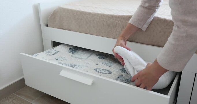 Woman takes folded blanket from under bed storage drawer. Minimalist organization brings neat order to bedroom space. Smart home storage solutions maximize small apartment functionality.