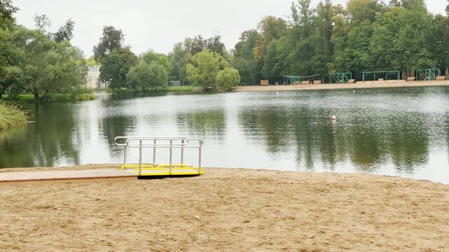 A quiet beach ramp extends toward a calm lake in Pushkin, Saint Petersburg. Green trees surround the peaceful water under an overcast sky. The scene inspires serene comfort and gentle relaxation.