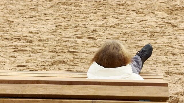 A woman rests comfortably on a wooden bench on a sandy beach in Pushkin, Saint Petersburg. She lies back quietly in full relaxation. Her peaceful moment feels calm and serene.