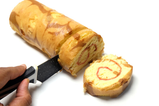 slicing a delicious yellow swiss roll cake with a red spiral jam layer using a black knife on a clean white background