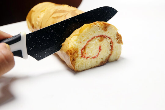 slicing a delicious yellow swiss roll cake with a red spiral jam layer using a black knife on a clean white background