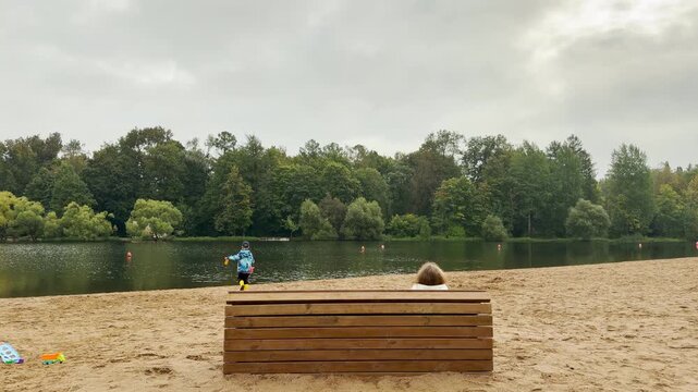 A woman rests on a bench while a child plays near the calm water in Pushkin, Saint Petersburg. Their quiet day brings a peaceful, serene feeling.