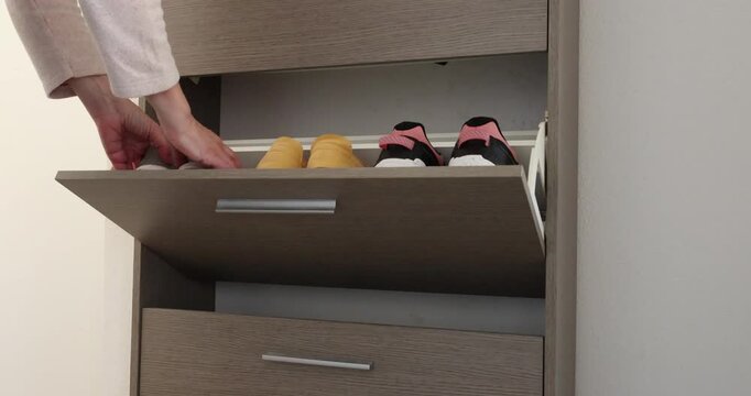 Woman puts grey sneakers into hallway shoe cabinet. Minimalist order creates tidy functional entryway atmosphere. Compact storage solutions inspire efficient small home organization.