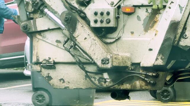A garbage truck mechanism lifts heavy trash bins on a city street. Workers operate the hydraulic machinery with steady focus. The essential routine brings a feeling of satisfaction and order.