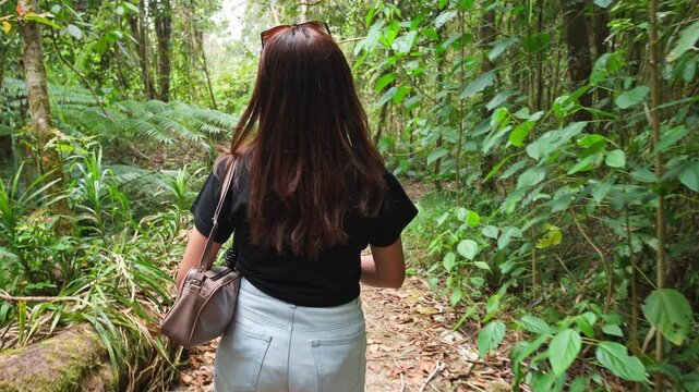 An Asian woman turns around and walks along a path surrounded by dense greenery in a tropical forest.
