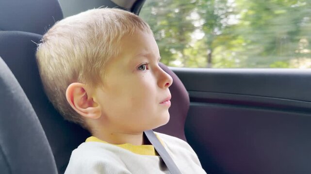 A young boy looks thoughtfully out the window while sitting in his car seat. He feels curious and calm as green trees blur past the moving vehicle. His quiet expression shows deep wonder.