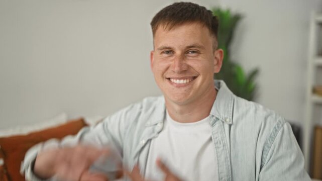 Man framing face with hands on bed in studio, seated among pillows and a plant, looking through hand frame at camera; playful curiosity.