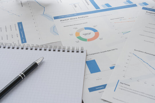 Blank grid notebook and pen on printed market share and project progress reports, with copy space.