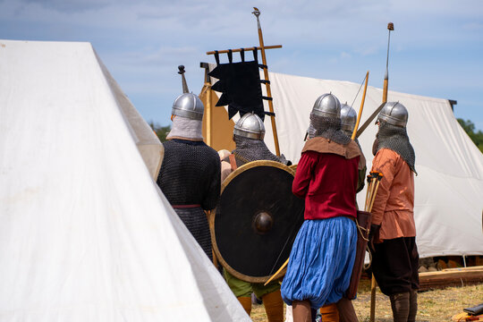 Medieval reenactors in helmets and chain mail gather among canvas tents at an outdoor historical camp, with a round shield, bows, and a black banner.