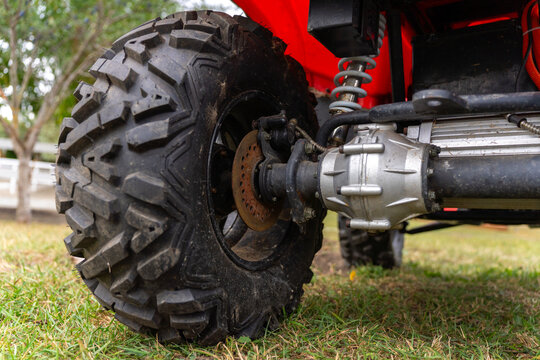 Close-up of a large knobby tire, exposed axle and silver differential housing beneath a red off-road vehicle, with a coil spring suspension above the grass.