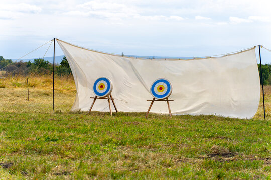 An outdoor archery practice range with two colorful bullseye targets on wooden stands in front of a large fabric backstop, set in a grassy field.