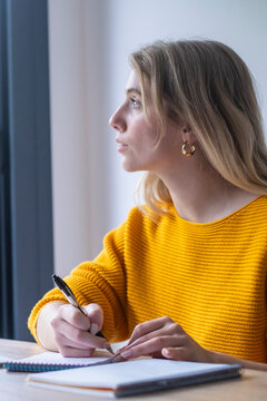 Young woman in a yellow sweater writing notes in a notebook with a pen, pausing to reflect and look out a window, capturing moments of inspiration and creative thought
