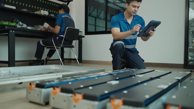 Technician wearing protective gloves services an electric vehicle battery pack in an auto workshop, inspecting modules and connections with tools beside a car.