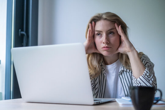 Young woman feeling overwhelmed and stressed while working on her laptop, experiencing a headache and symptoms of burnout from prolonged screen time and work pressure in a home office setting