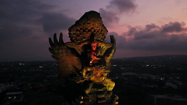 Illuminated Garuda Wisnu Kencana statue glowing at sunset dusk with dramatic twilight sky in Bali cultural park orbit aerial drone footage