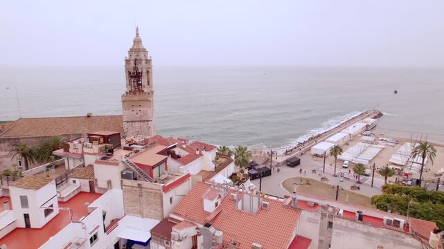 4K drone aerial view of Sitges, Spain, featuring the iconic Sant Bartomeu i Santa Tecla church overlooking the Mediterranean coast, beaches and historic seaside town.