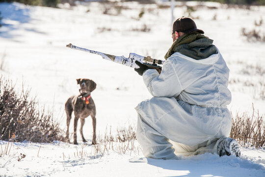 Hunter in snow camouflage crouches with his rifle and dog
