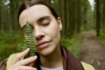 Close up of young adult Caucasian woman holding green fern leaf near face with eyes closed in forest. Visual representing natural skincare and mental wellbeing campaigns © Seventyfour