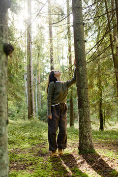 Vertical shot of young adult Caucasian woman touching tree trunk in forest, looking up. Visual concept for environmental awareness, ecotourism, mental wellness campaign