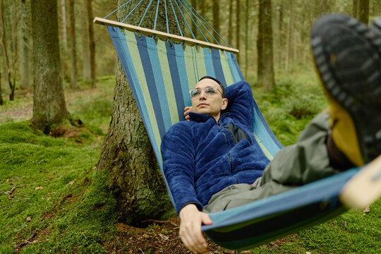 Young adult Caucasian man relaxing in hammock between trees in forest, enjoying peaceful recreation. Suitable for outdoor lifestyle, mental wellness and ecotourism campaigns