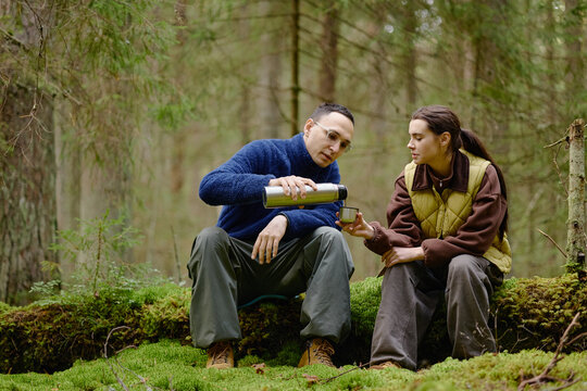 Young adult Caucasian man pouring hot drink from thermos to young adult Caucasian woman while resting on mossy log in forest. Scene suitable for outdoor recreation camping campaigns
