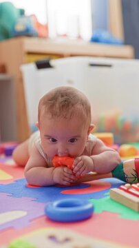 Baby boy mouthing orange teether while grasping it with chubby hand on colorful playmat in a home playroom; curiosity.